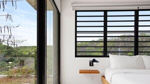 A bedroom with a large window, a bed, a nightstand, and a view of greenery outside.