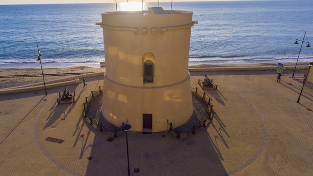 Un bâtiment circulaire doté d’une porte centrale, entouré d’une zone pavée avec des bancs et des lampadaires, et offrant une vue sur la mer.