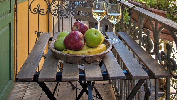 Una mesa de madera con un frutero y copas de vino en un balcón con barandillas de hierro forjado.