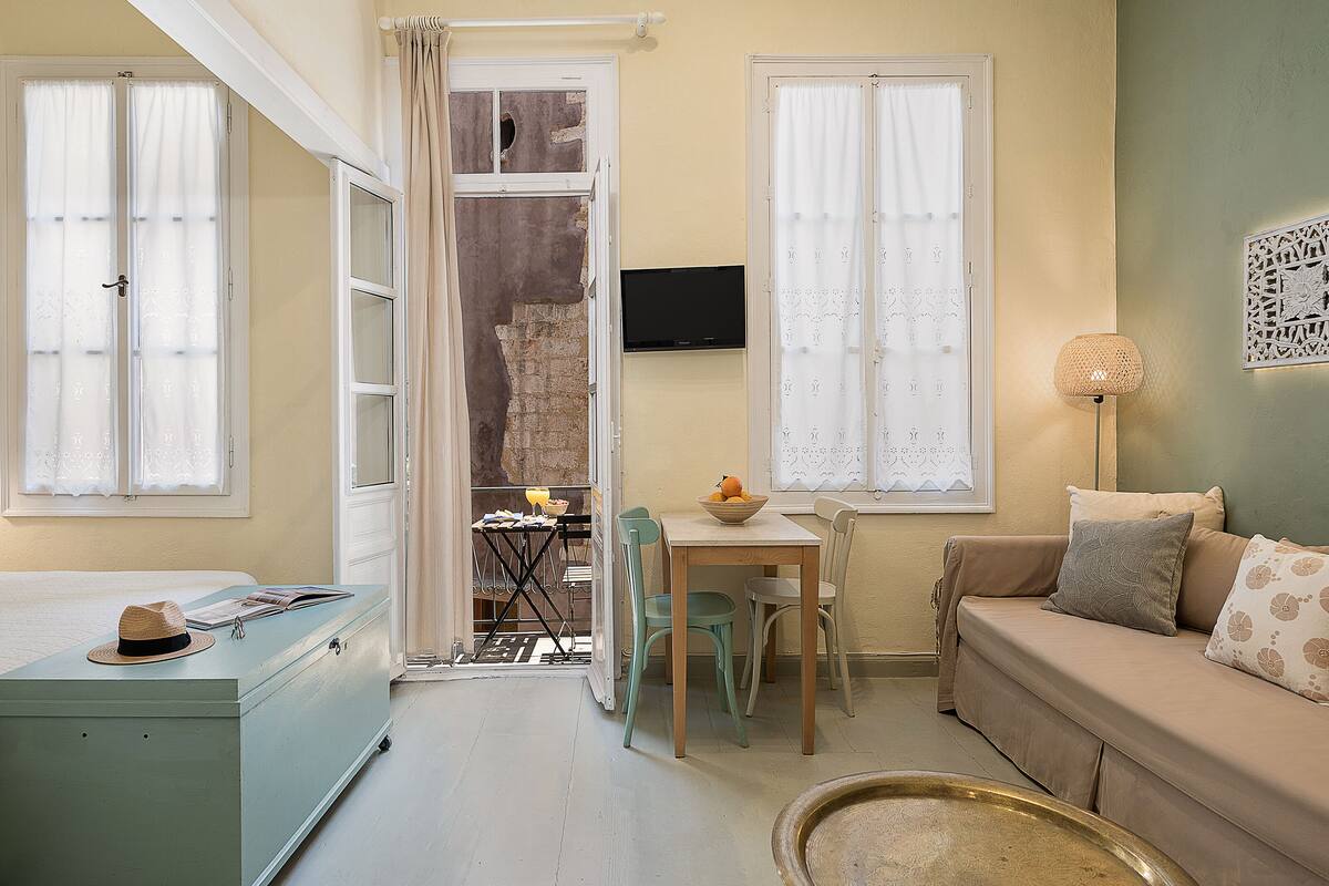 Une pièce confortable avec un canapé, une table à manger et des chaises, un téléviseur fixé au mur et une valise posée sur le sol.