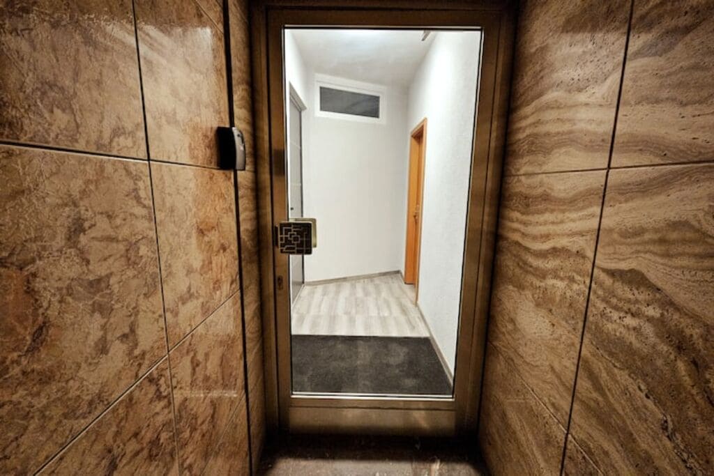 Un corridoio stretto con una porta che dà su una stanza. Il corridoio presenta pavimento e pareti piastrellate, e una piccola finestra sopra la porta.