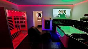 A modern indoor pool area with a jacuzzi, a lounge chair, and a large screen displaying a 'SUITS' advertisement. - Magnifique Suite Spa Luxembourg (Differdange)