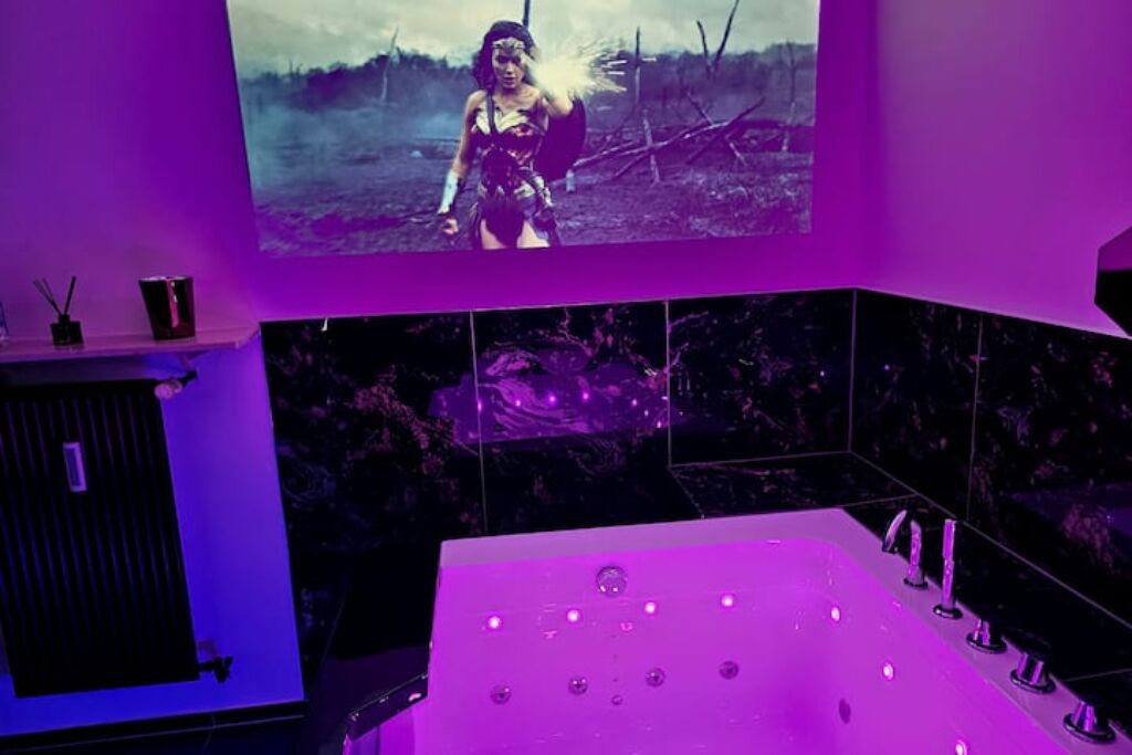 Ett rum med en stor, platt-TV som är monterad på väggen, ett vitt badkar med flera massagemunstycken och en väggbeklädnad som liknar marmor.