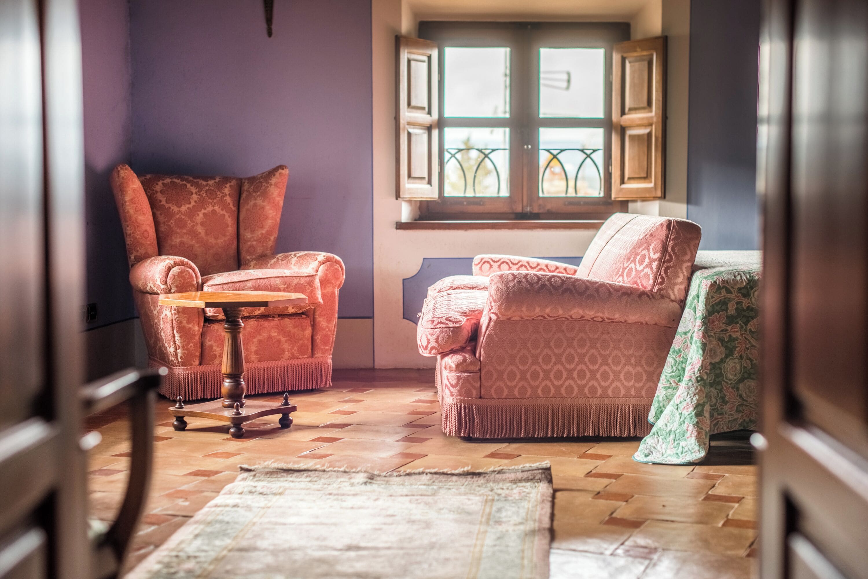 Una habitación acogedora con un sillón estampado, un sofá a juego y una mesita pequeña. Se ve una ventana con contraventanas, y una alfombra estampada cubre el suelo.