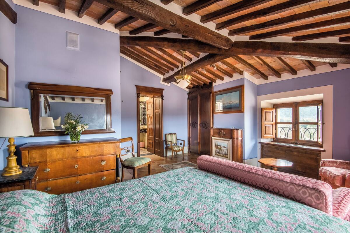 Une chambre avec un plafond en bois, un lit recouvert d’une couvre-lit à motifs floraux, une commode, une chaise, une petite table et une fenêtre donnant sur un beau paysage.
