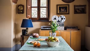 Una taula de menjador amb un bol de fruita, un plat amb pastissos i una làmpada. Hi ha reproductors de discos antics en un moble, un rellotge a la paret i una finestra amb vista a vegetació.