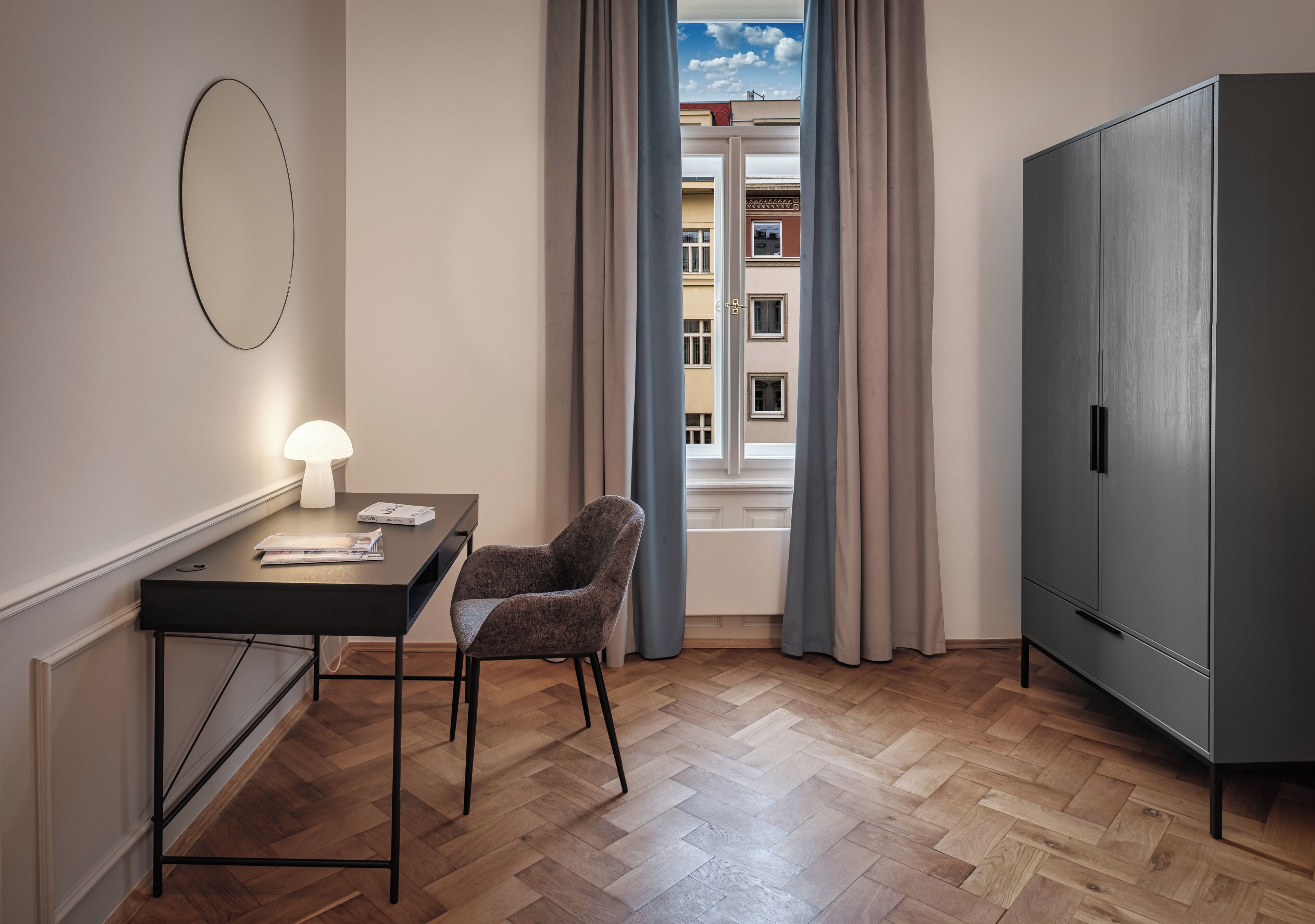 A modern room with a wooden herringbone floor, a desk with a lamp, a chair, and a wardrobe.