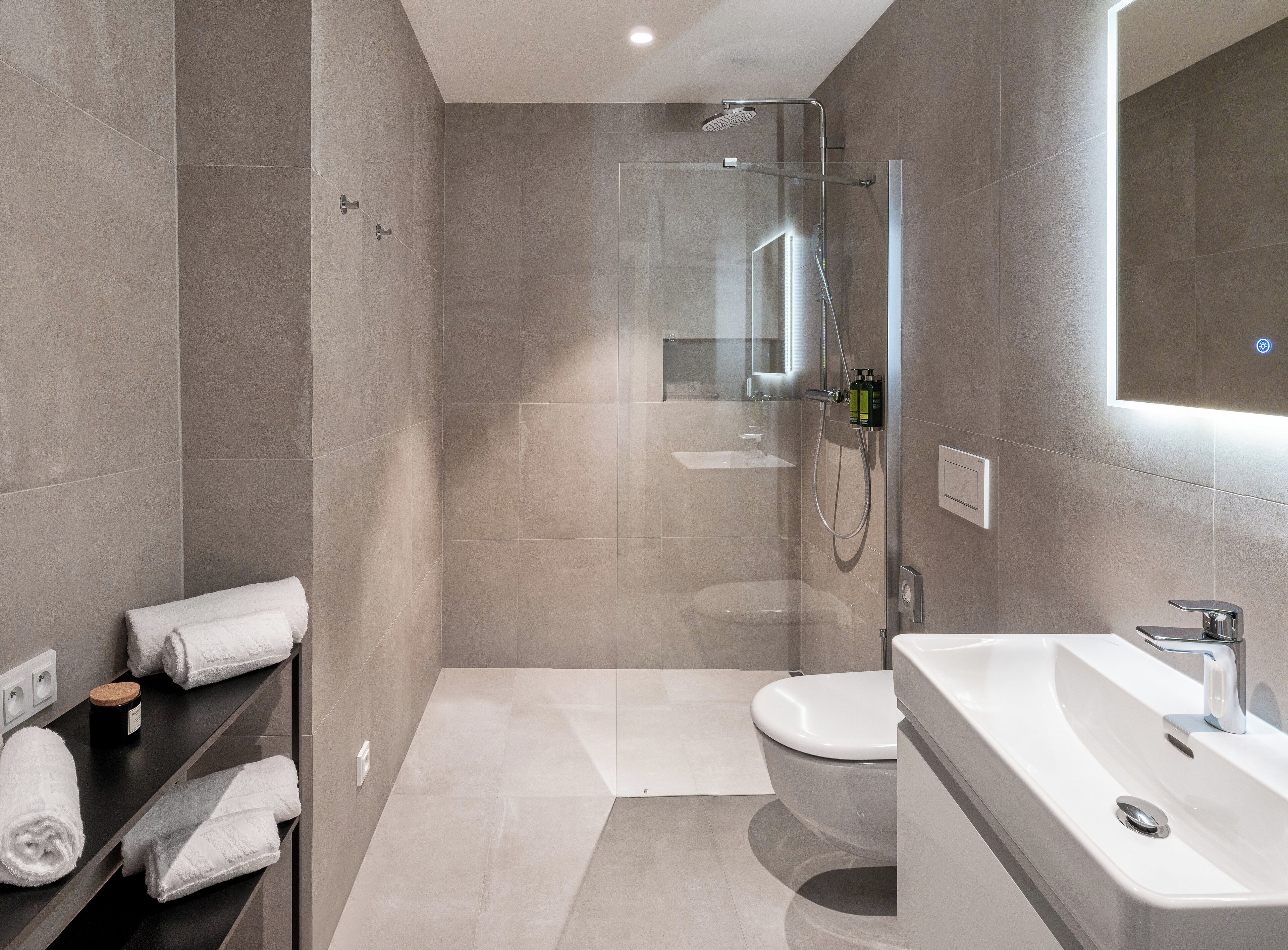 A modern bathroom with a glass-enclosed shower, a white sink, and a toilet. There are towels on a shelf and a small bottle of shampoo.