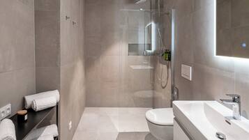 A modern bathroom with a glass-enclosed shower, a white sink, and a toilet. There are towels on a shelf and a small bottle of shampoo.