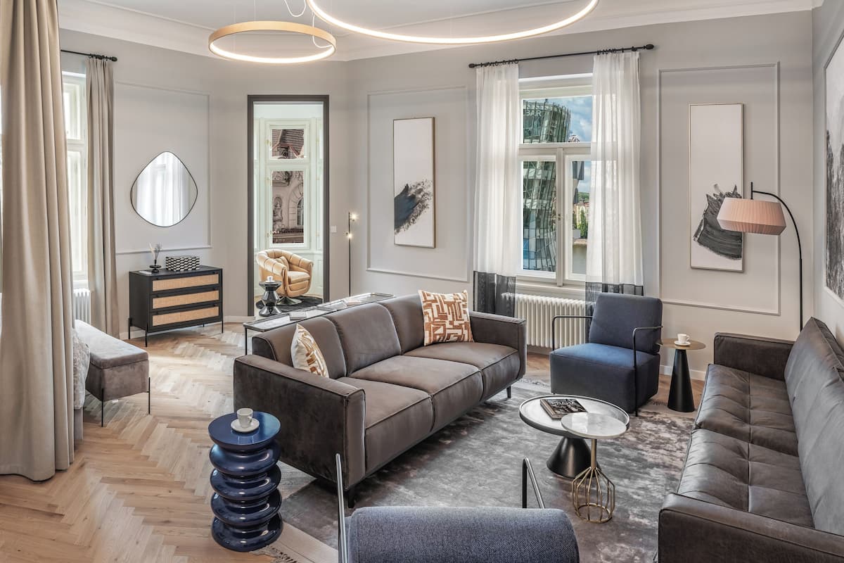 A modern living room with a sofa, armchairs, a coffee table, and a side table. There is a window with curtains, a radiator, and a mirror on the wall.