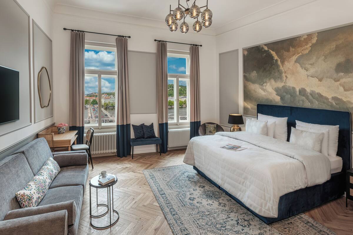 A spacious bedroom with a large bed, a sofa, a small table, and a view of the city through large windows.