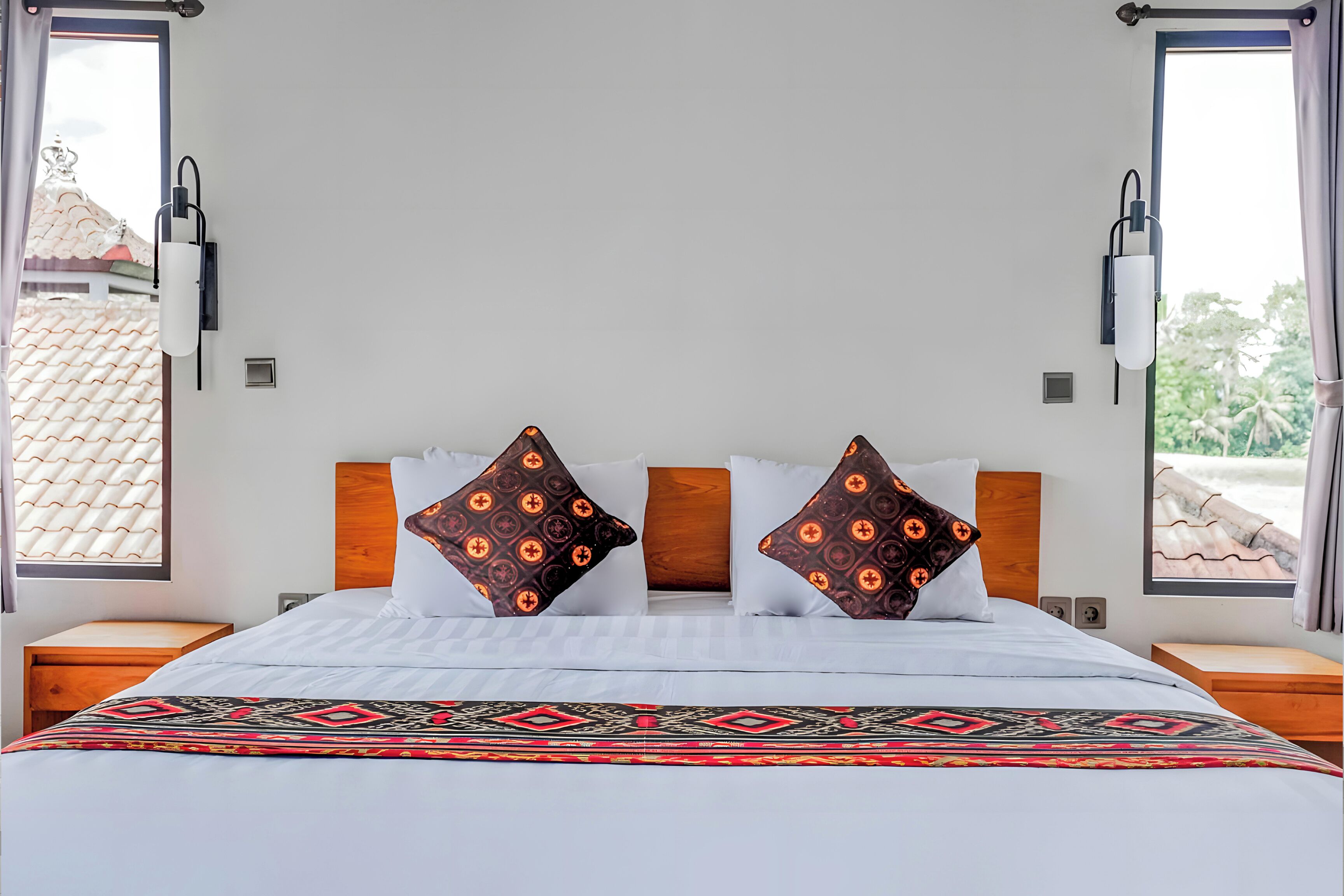 A hotel room with a large bed, two nightstands, a wooden headboard, and a view of a tiled roof and greenery outside.