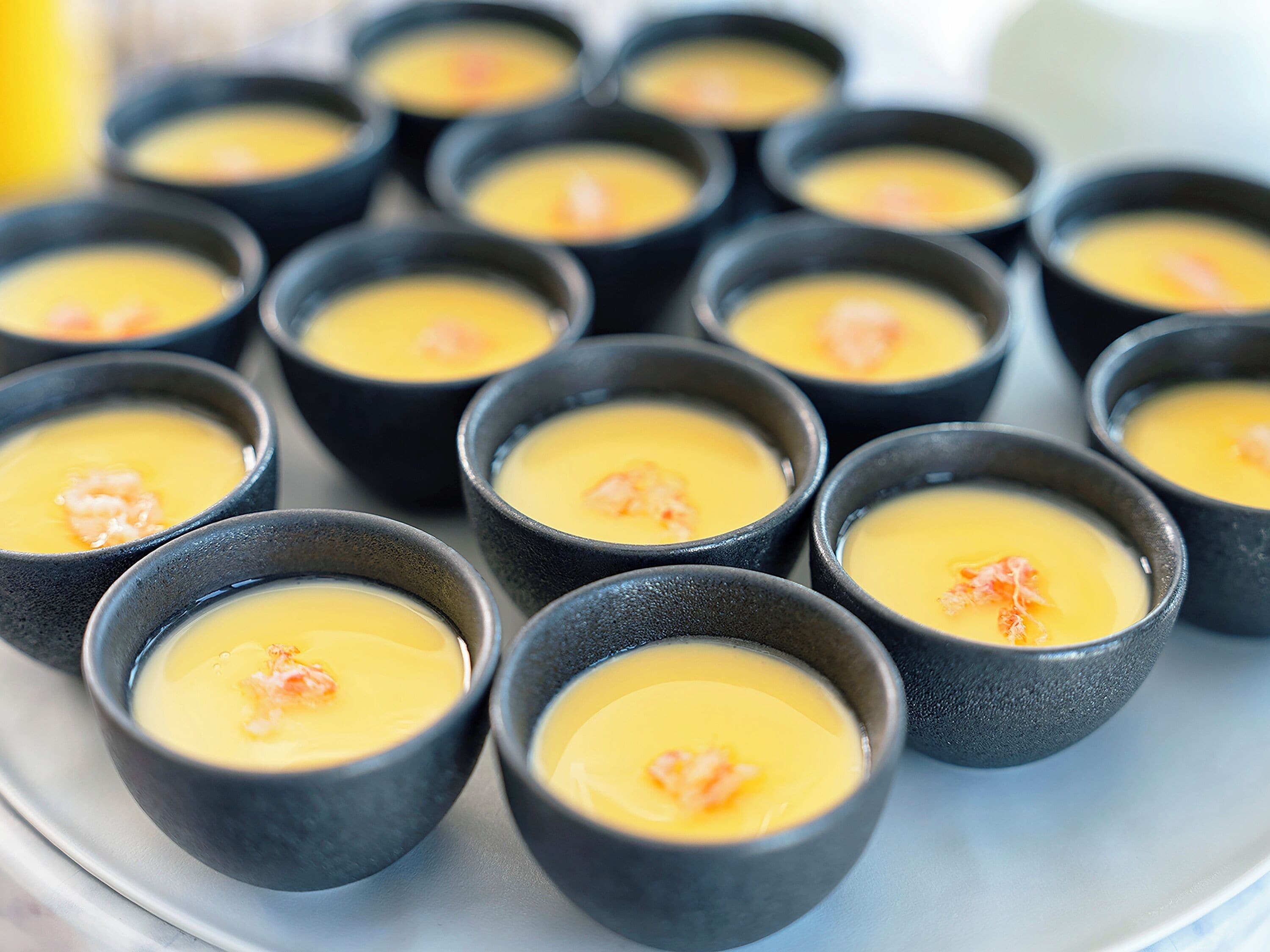 A series of small bowls with a creamy yellow dessert topped with a pinkish ingredient, possibly shrimp, arranged on a light-colored surface.