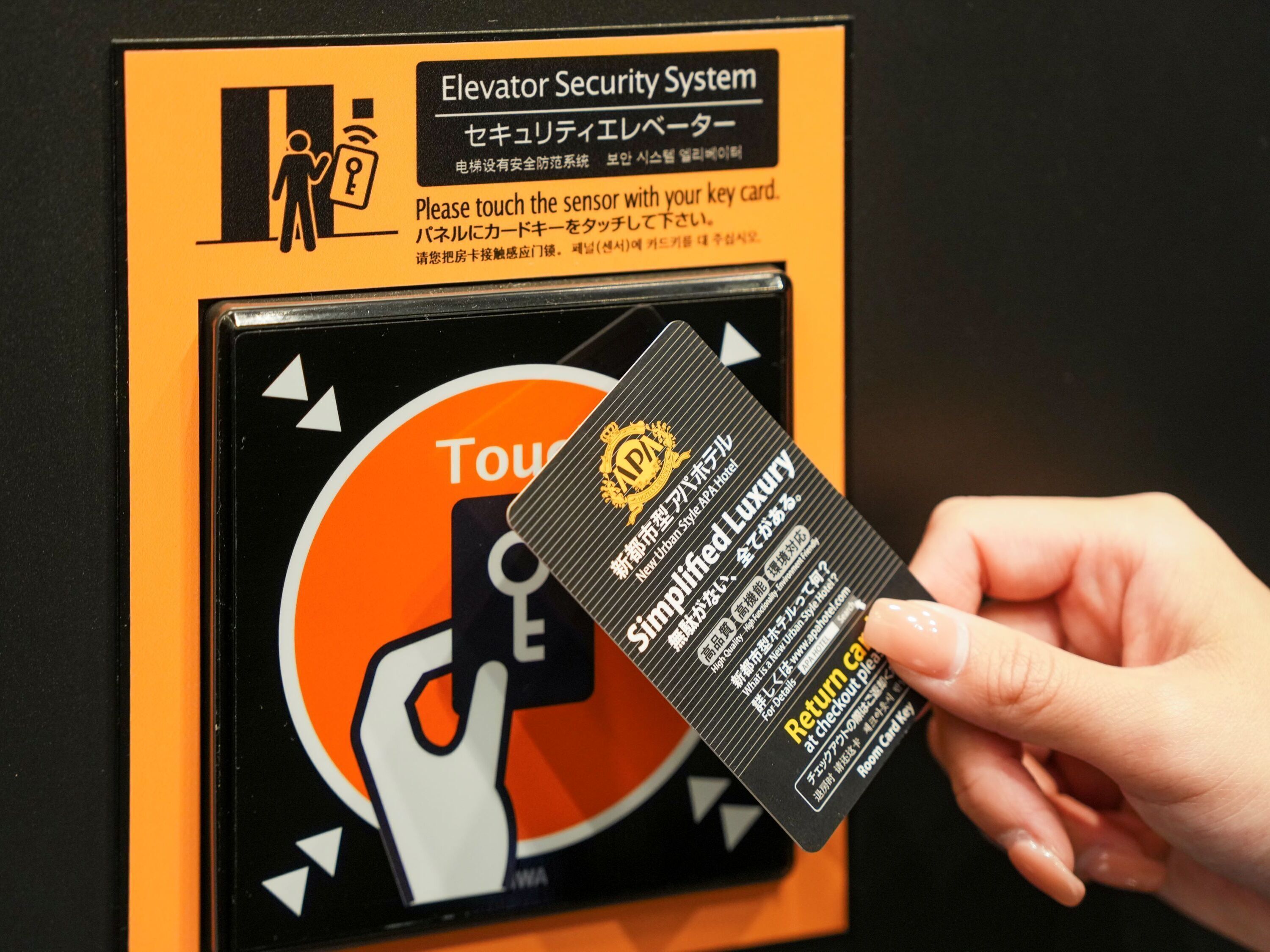 Une main tenant une carte à puce avec du texte et un symbole, devant un panneau indiquant le système de sécurité d’un ascenseur.