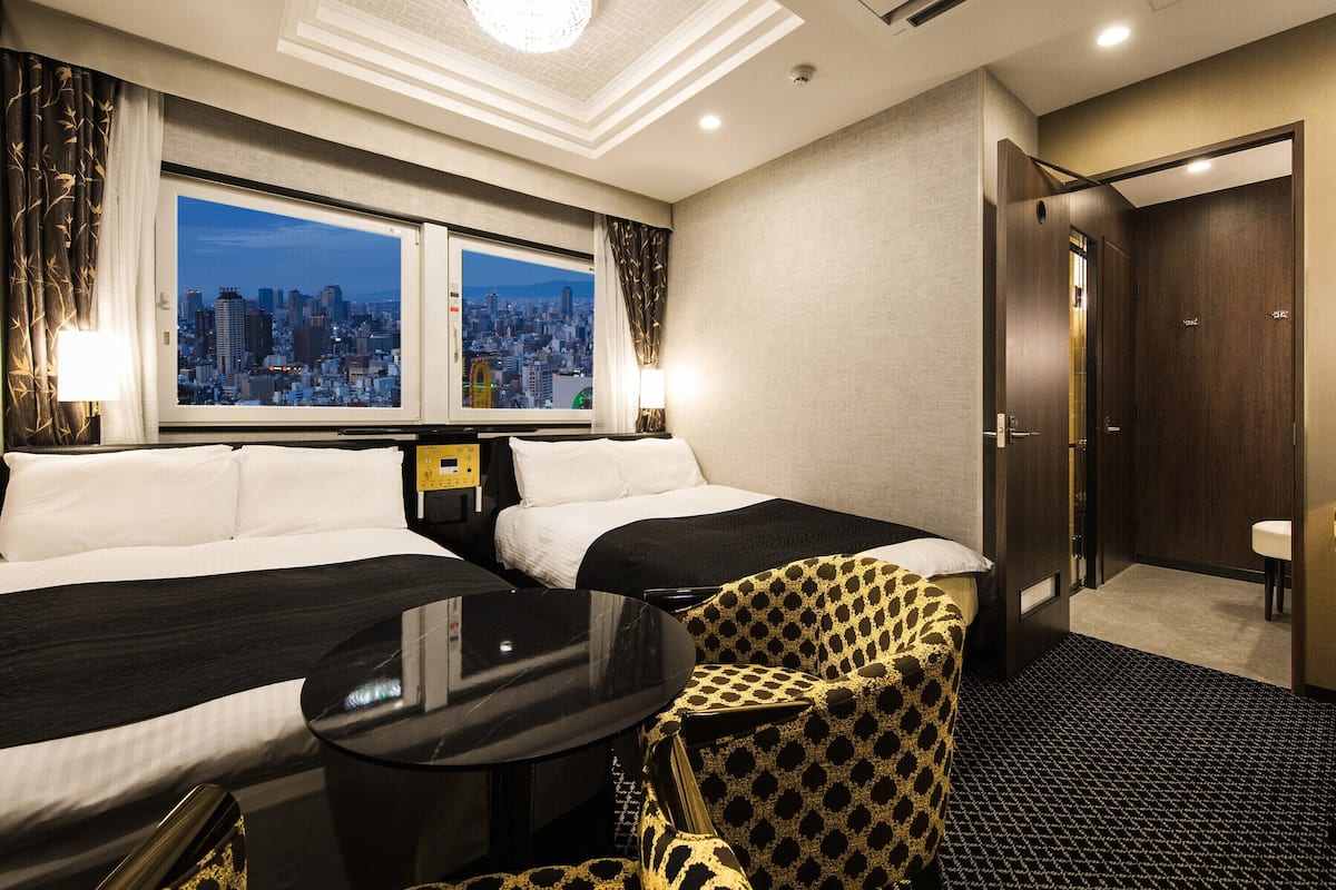 Een hotelkamer met twee bedden, een klein tafeltje, een stoel, en uitzicht op de stad, plus een deur.