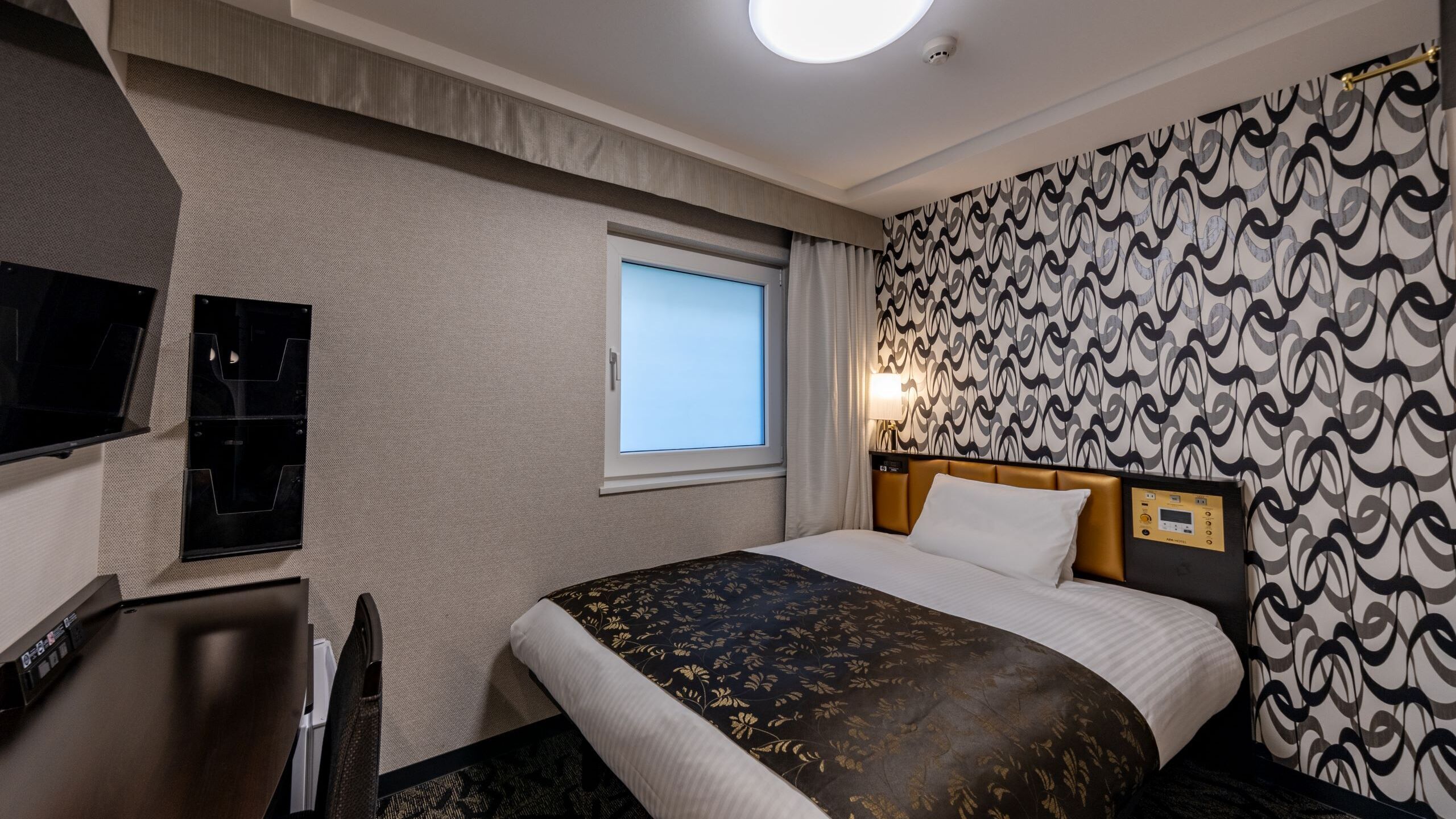 Une chambre d’hôtel avec un lit, un bureau, une télévision, une fenêtre et un papier peint à motifs.