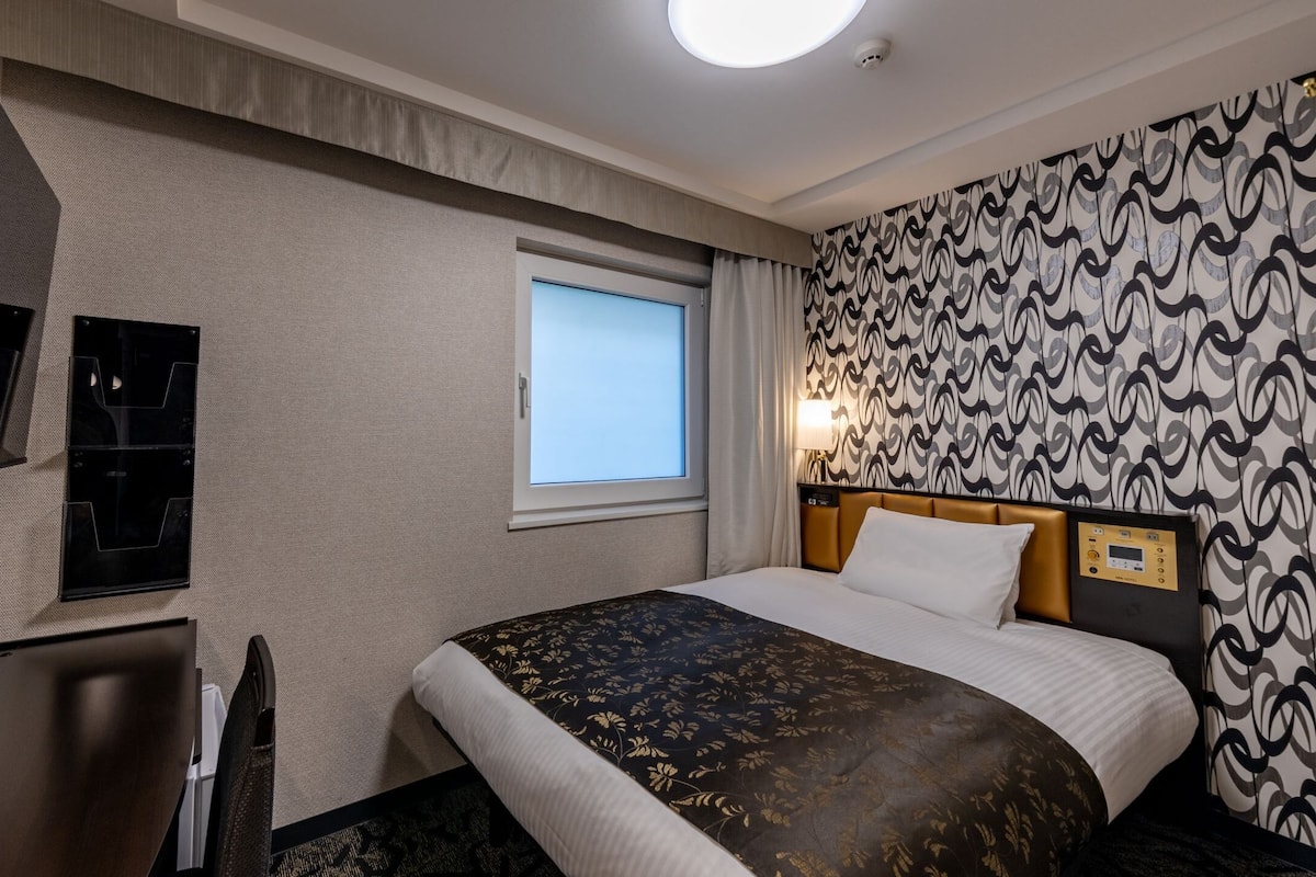 Een hotelkamer met een bed, een bureau, een televisie, een raam en behang met een patroon.