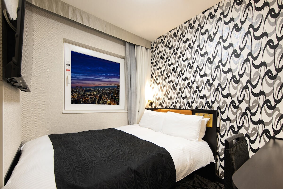 Hotelkamer met een bed, uitzicht op een stadsbeeld, een behang met patroon en een klein tafeltje.
