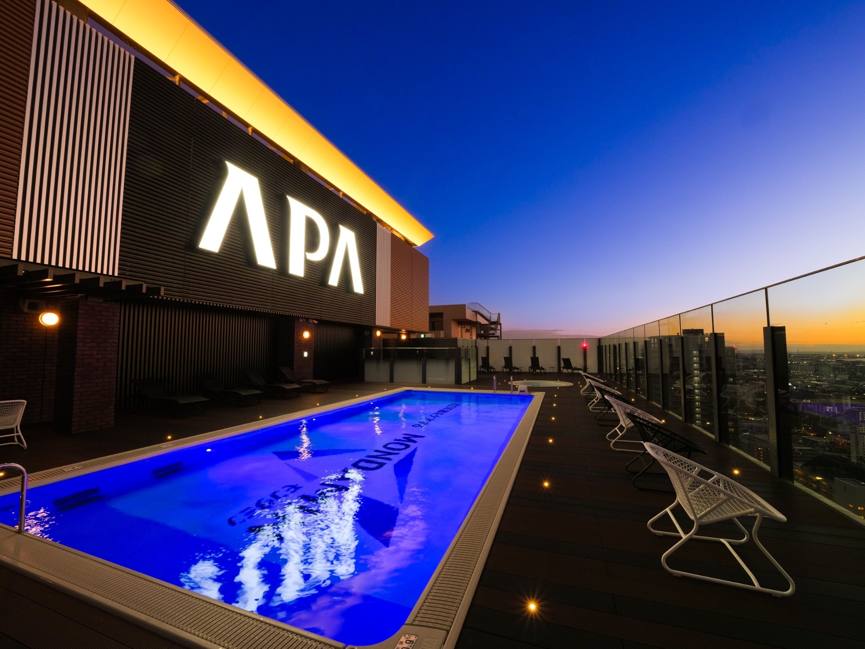 A rooftop pool with a view of the city at dusk, a large illuminated sign reading "APA", and lounge chairs along the poolside.