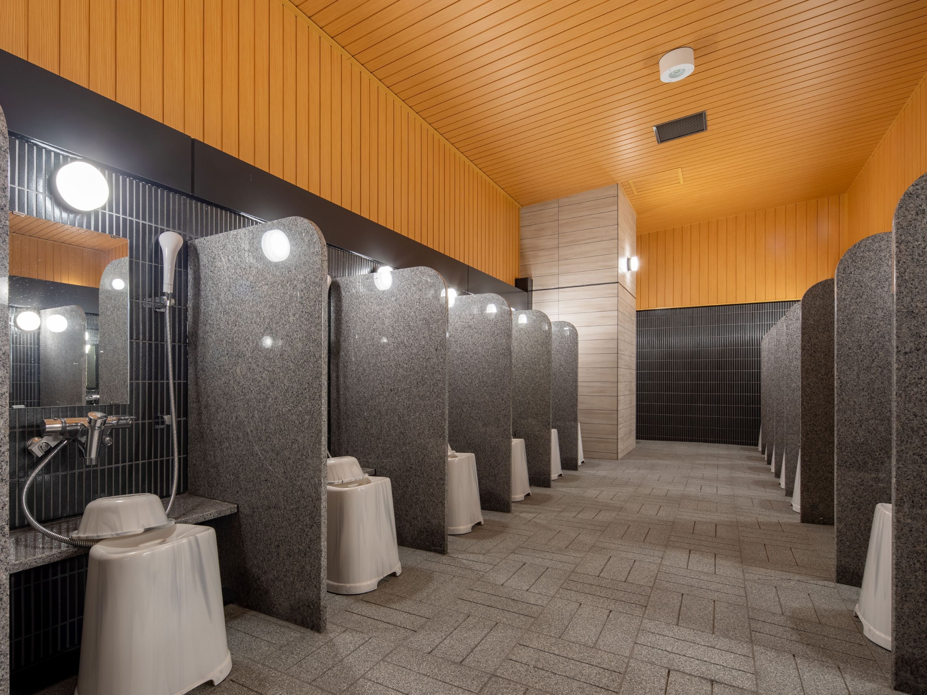 A row of modern, granite-finished public restrooms with individual sinks and mirrors, wooden paneling on the ceiling, and a tiled floor.