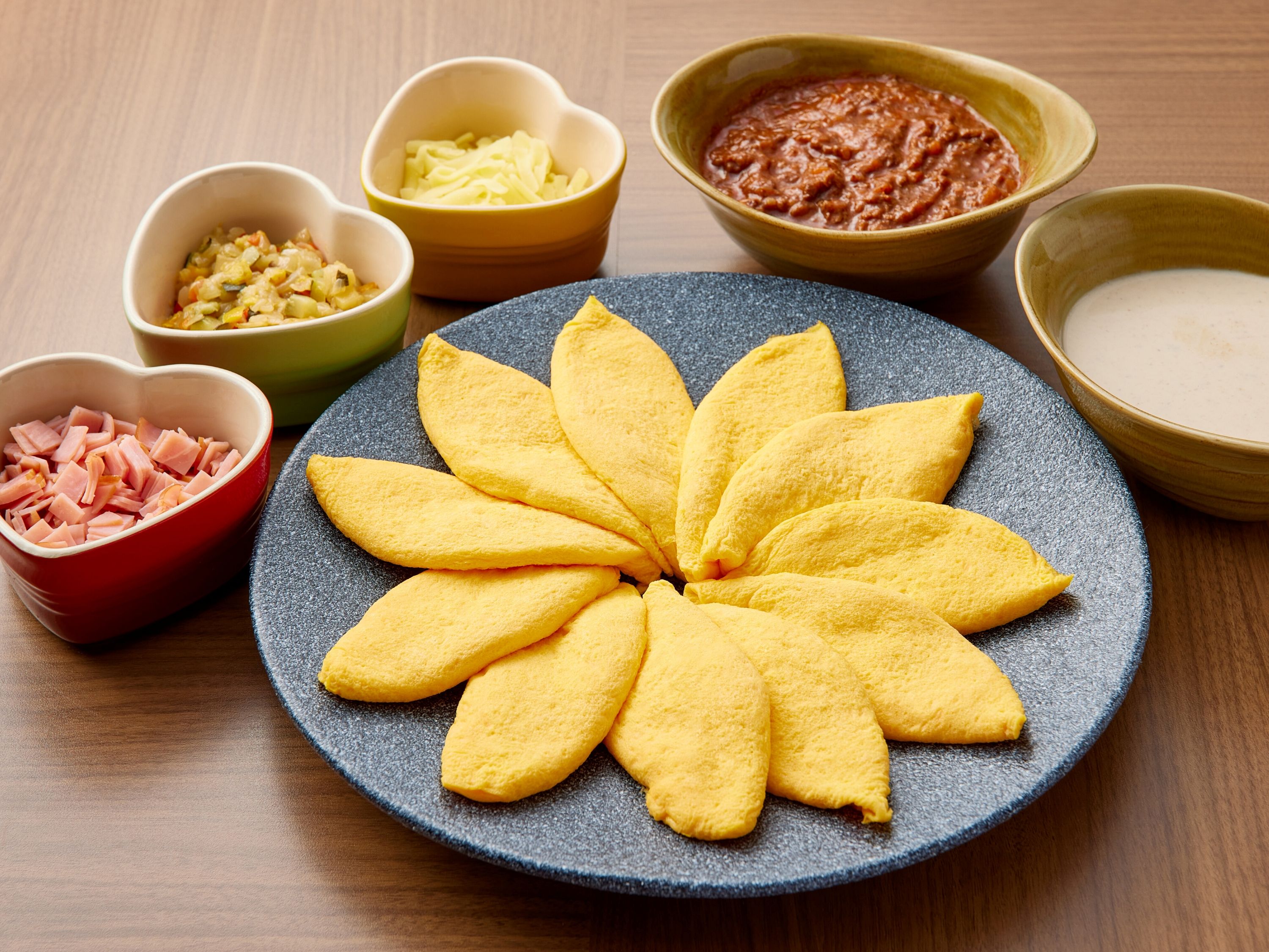 A plate of yellow pastries arranged in a flower pattern, surrounded by small bowls containing various ingredients.