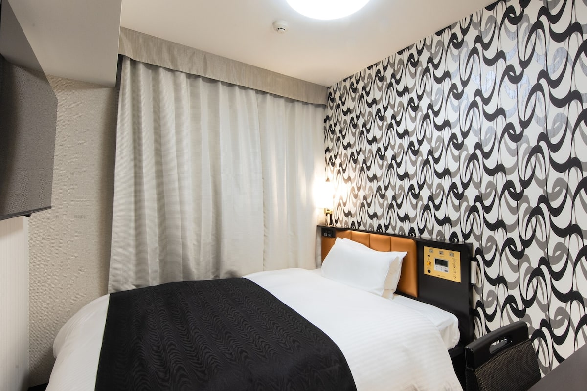 Hotelkamer met een bed, een hoofdbord met bedieningselementen, een muur met behang met patroon en een raam met gordijnen.
