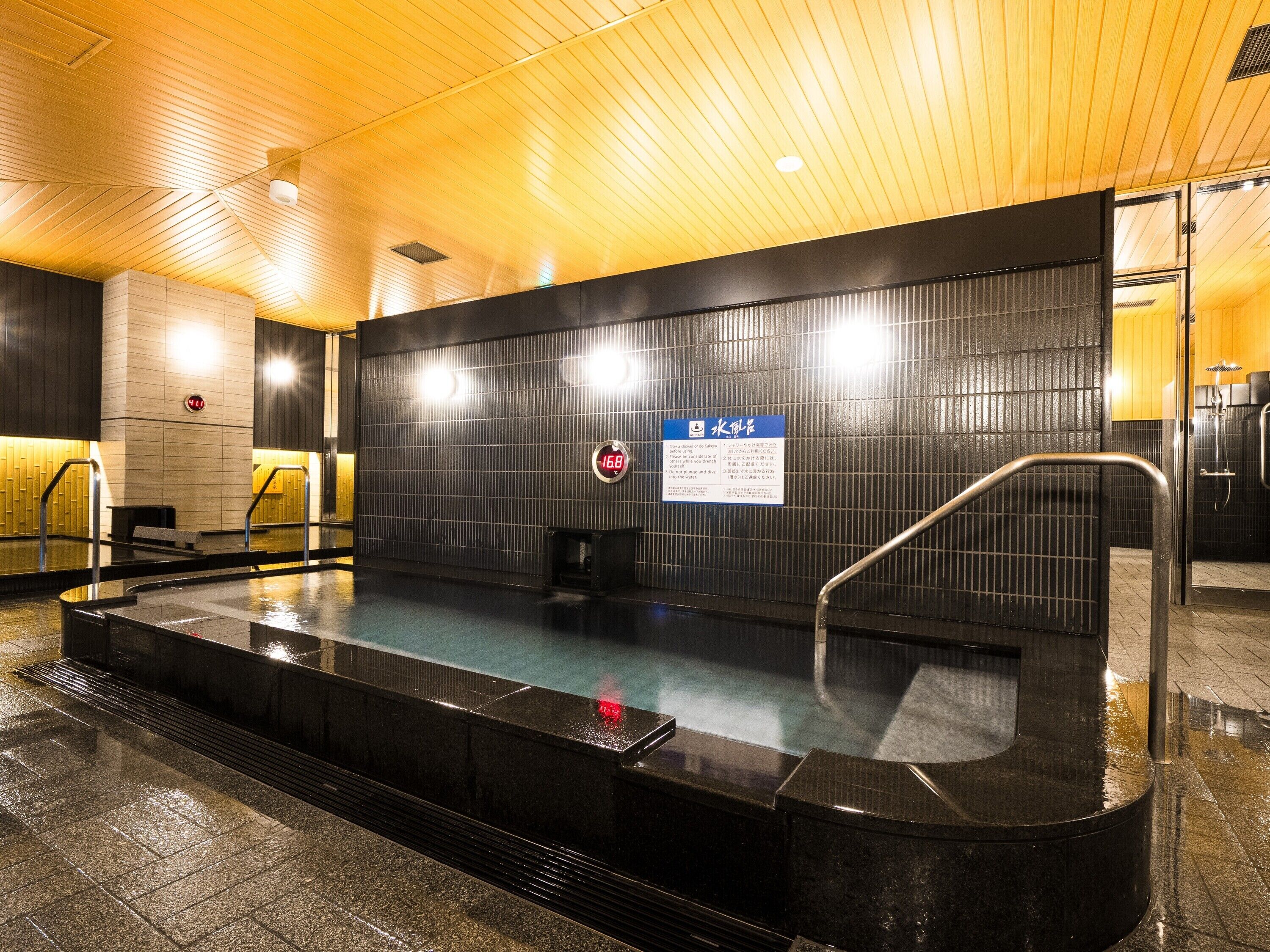 Ein öffentliches Hallenbad mit gefliestem Boden, schwarzem Beckenrand und einem Schild mit japanischem Text.