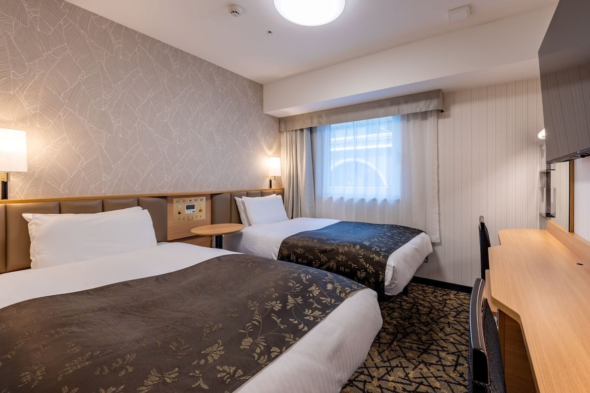 Een hotelkamer met twee bedden, een bureau, nachtkastje, lamp en een raam met gordijnen.