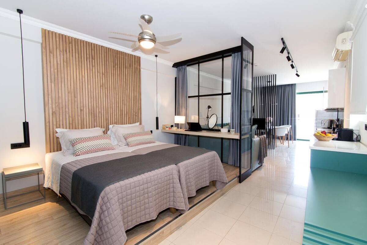 A modern hotel room with a large bed, a desk, a ceiling fan, and a view of the kitchen.