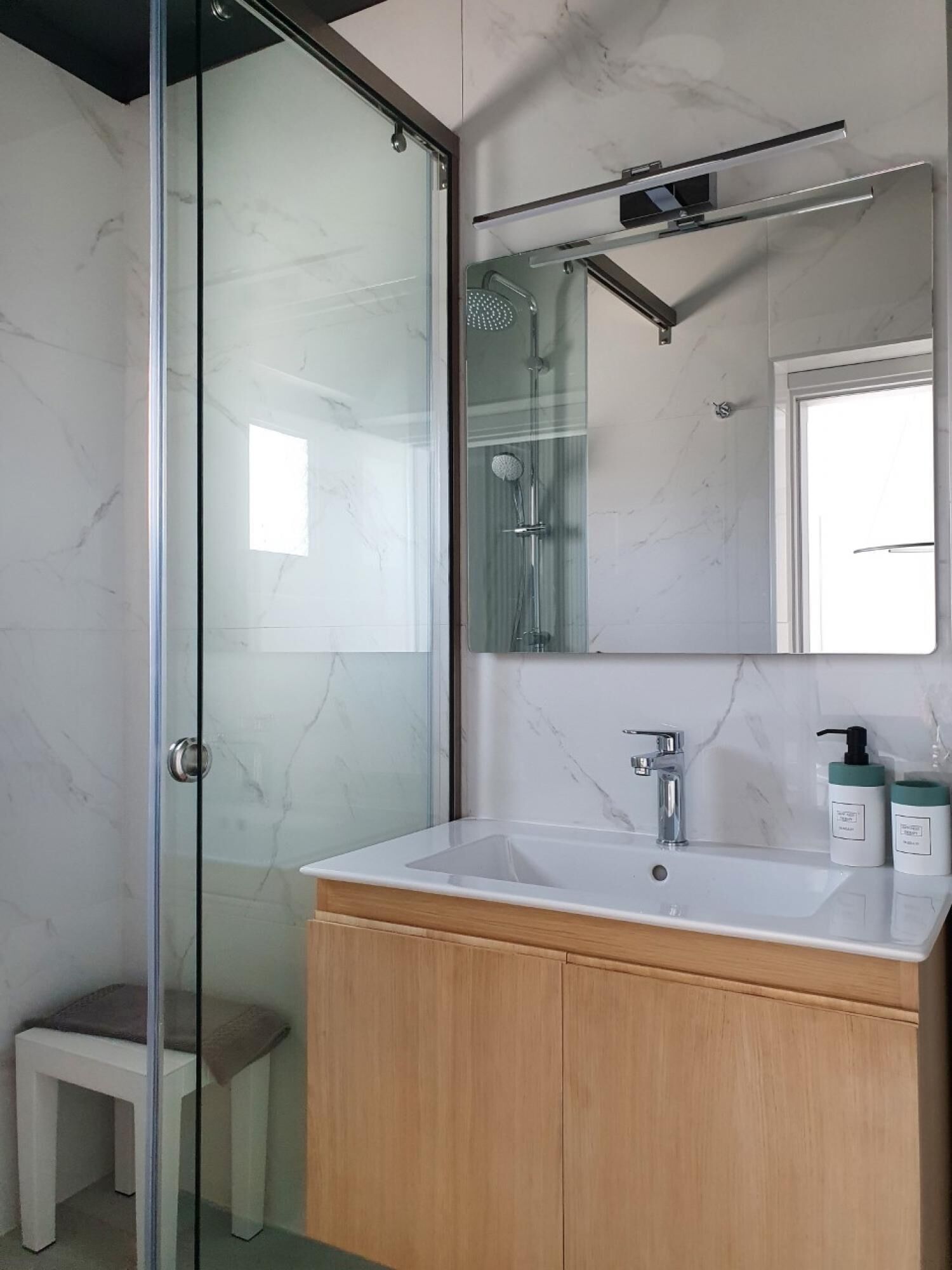 A modern bathroom with a glass-enclosed shower, a wooden vanity, and a marble-tiled wall.