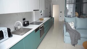 A modern kitchen with white cabinets, a built-in oven, and a large island with a bowl. There is a light blue sofa with a blanket and pillows in the living area. - Phaedra's Studios in Kolymbari (Platanias)