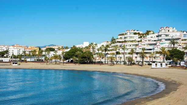 A coastal hotel with multiple floors, palm trees, and a beach in the foreground.