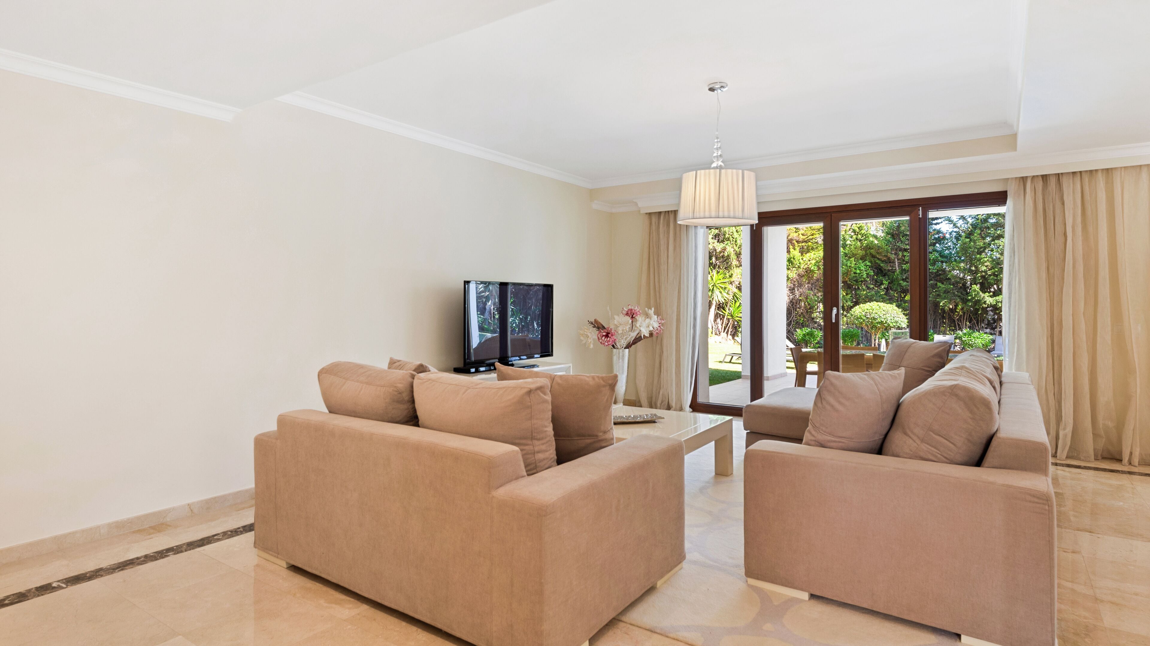 Una sala de estar moderna con sofás color beige, un televisor de pantalla plana y vistas a un jardín a través de amplios ventanales.