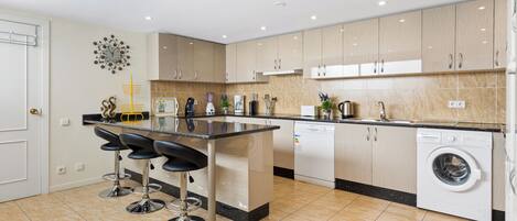 A modern kitchen with a central island, bar stools, a washing machine, and a sink.