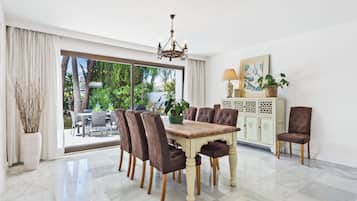 A dining area with a wooden table and chairs, a chandelier, a cabinet with decorative panels, and a view of greenery outside.