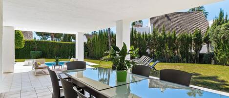 A covered outdoor dining area with a pool, lounge chairs, and a potted plant.