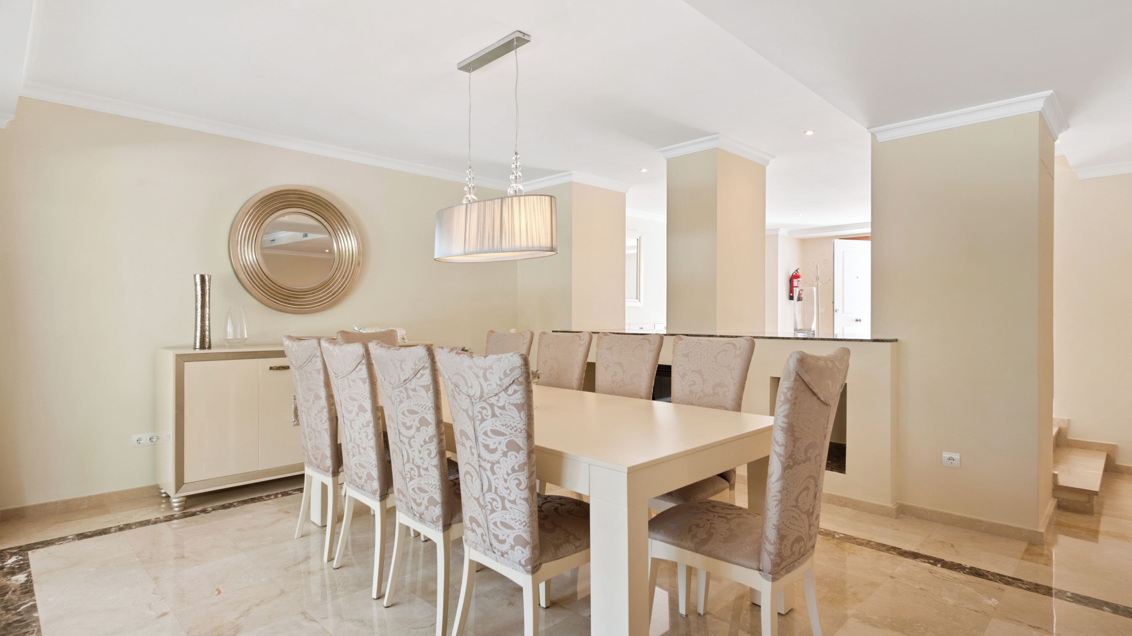 A dining area with a light-colored table and patterned chairs, a modern pendant light, and a decorative mirror on the wall.