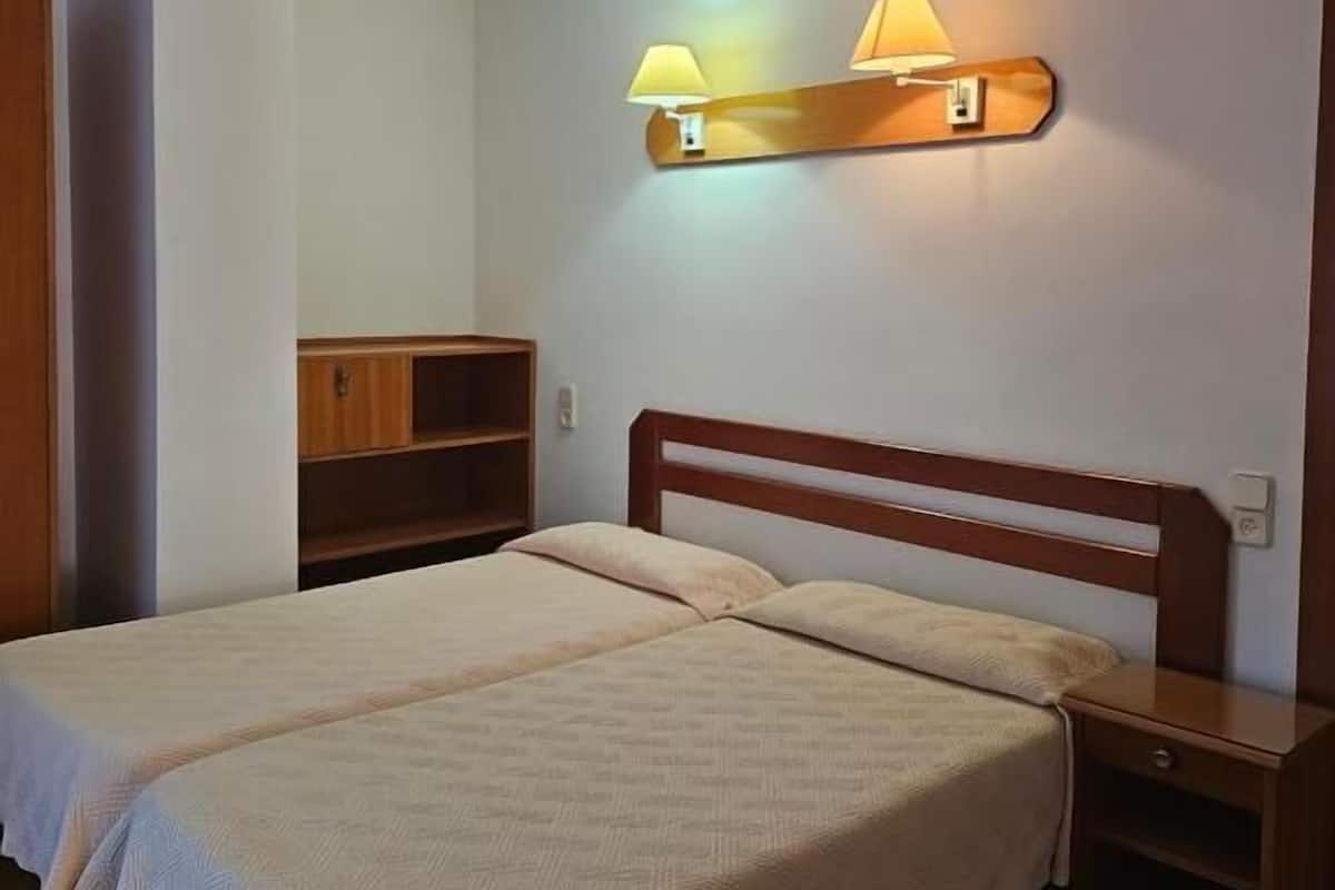 Una habitación con dos camas, cabeceros de madera, una mesita de noche, un estante y una lámpara de pared.