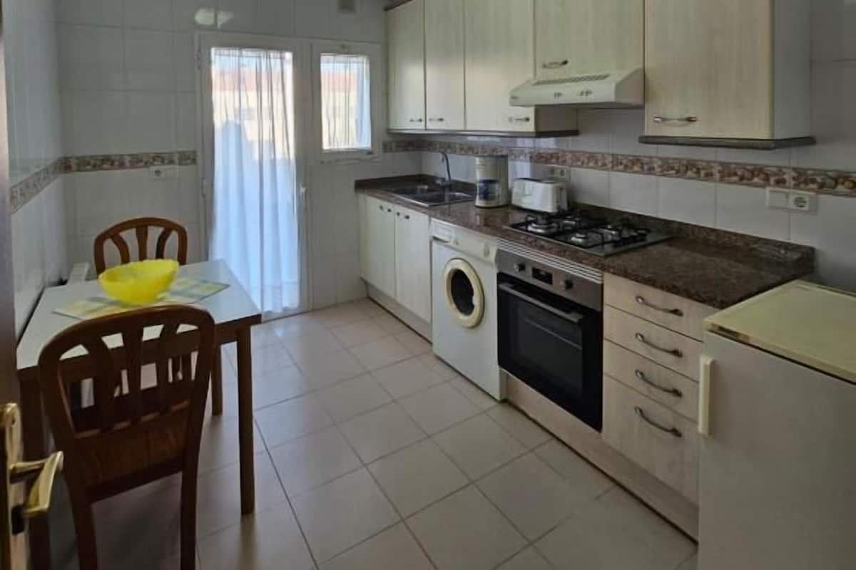 Cocina compacta con armarios blancos, lavadora, estufa y comedor con mesa y sillas.