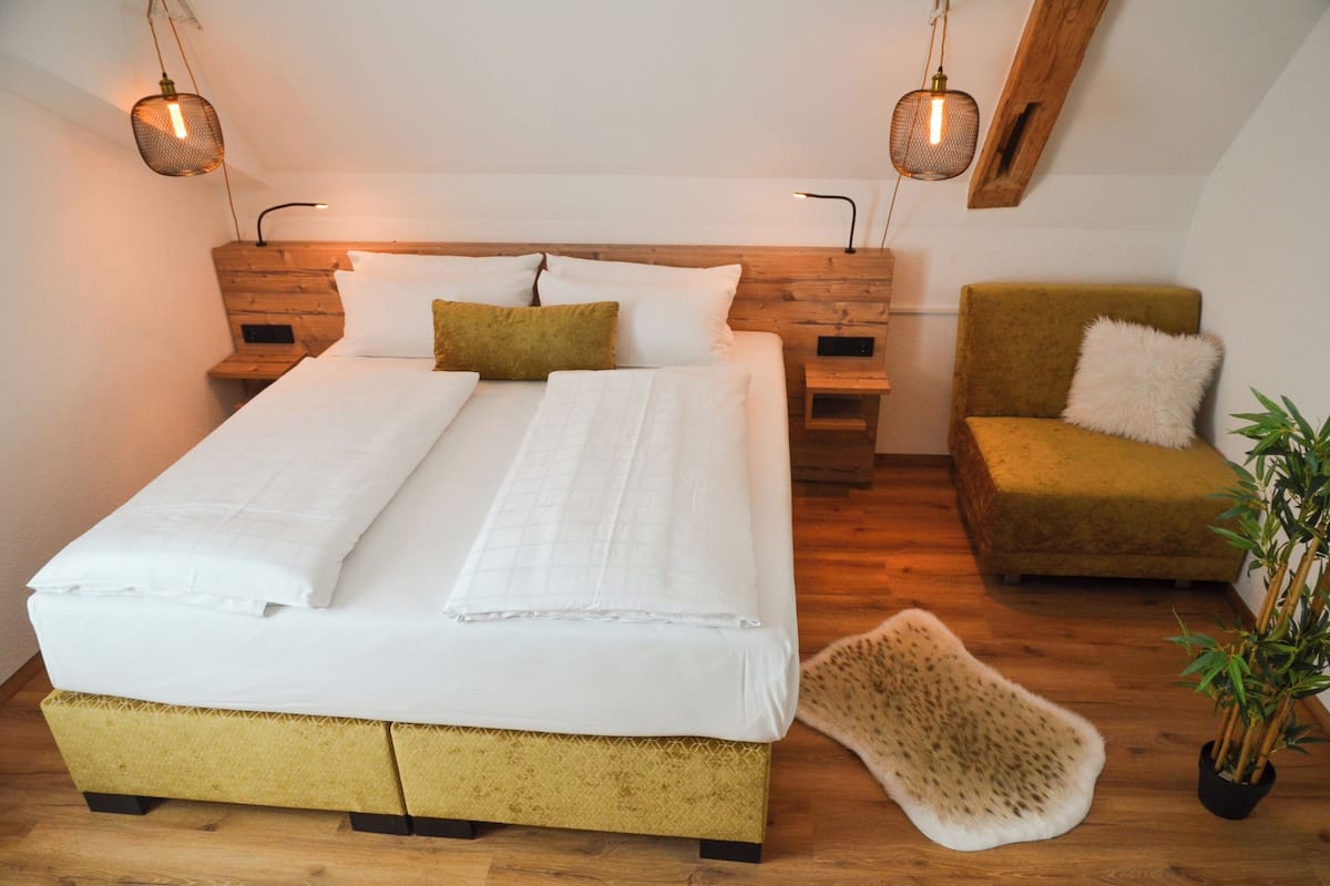 A double bed with white linens, a wooden headboard, two bedside tables, a green armchair with a white pillow, and a small potted plant.