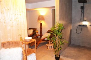 A spa room with a wooden chair, a small table with a white bowl, a potted plant, and a wall-mounted shower.