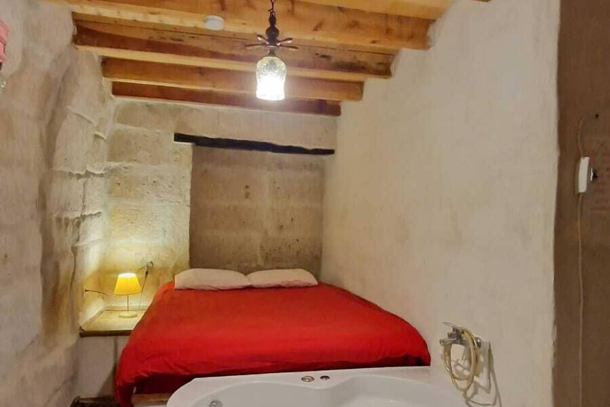Una habitación pequeña y rústica con techo de madera, una cama individual con manta roja, una bañera blanca grande y una mesita con una lámpara.