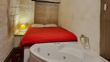A small, rustic room with a wooden ceiling, a single bed with a red blanket, a large white bathtub, and a small table with a lamp.