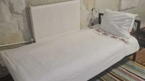A single bed with a white mattress and pillow, a white radiator, and a colorful striped rug on a concrete floor. - King Solomon Palace  (Avanos)
