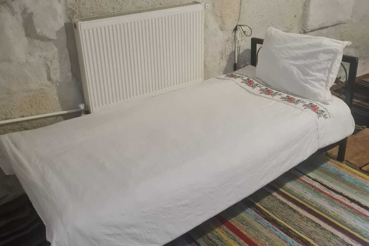 Una cama individual con colchón y almohada blancos, un radiador blanco y una alfombra colorida a rayas sobre un piso de concreto.