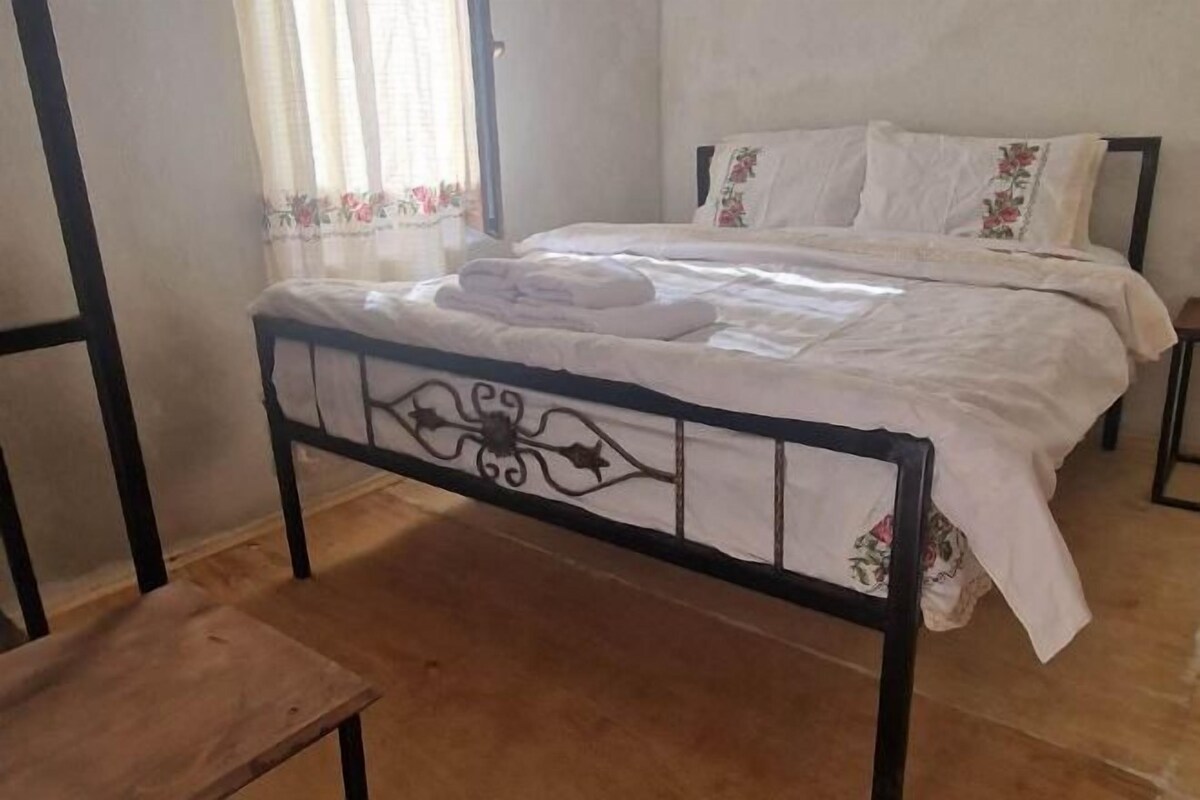 Una cama individual con estructura metálica y ropa de cama blanca, un banco de madera y una ventana con cortinas florales.