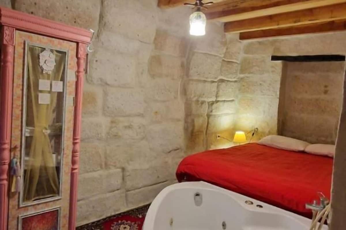 Una habitación con paredes de piedra, una cama, una manta roja, una bañera blanca y un espejo con marco rosa.