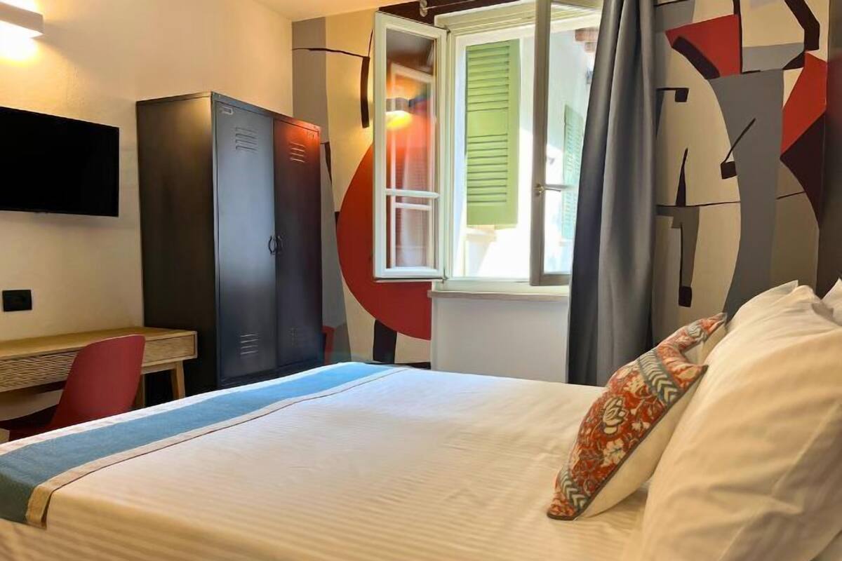 Quarto de hotel com cama, escrivaninha, guarda-roupa e janela com persianas verdes.