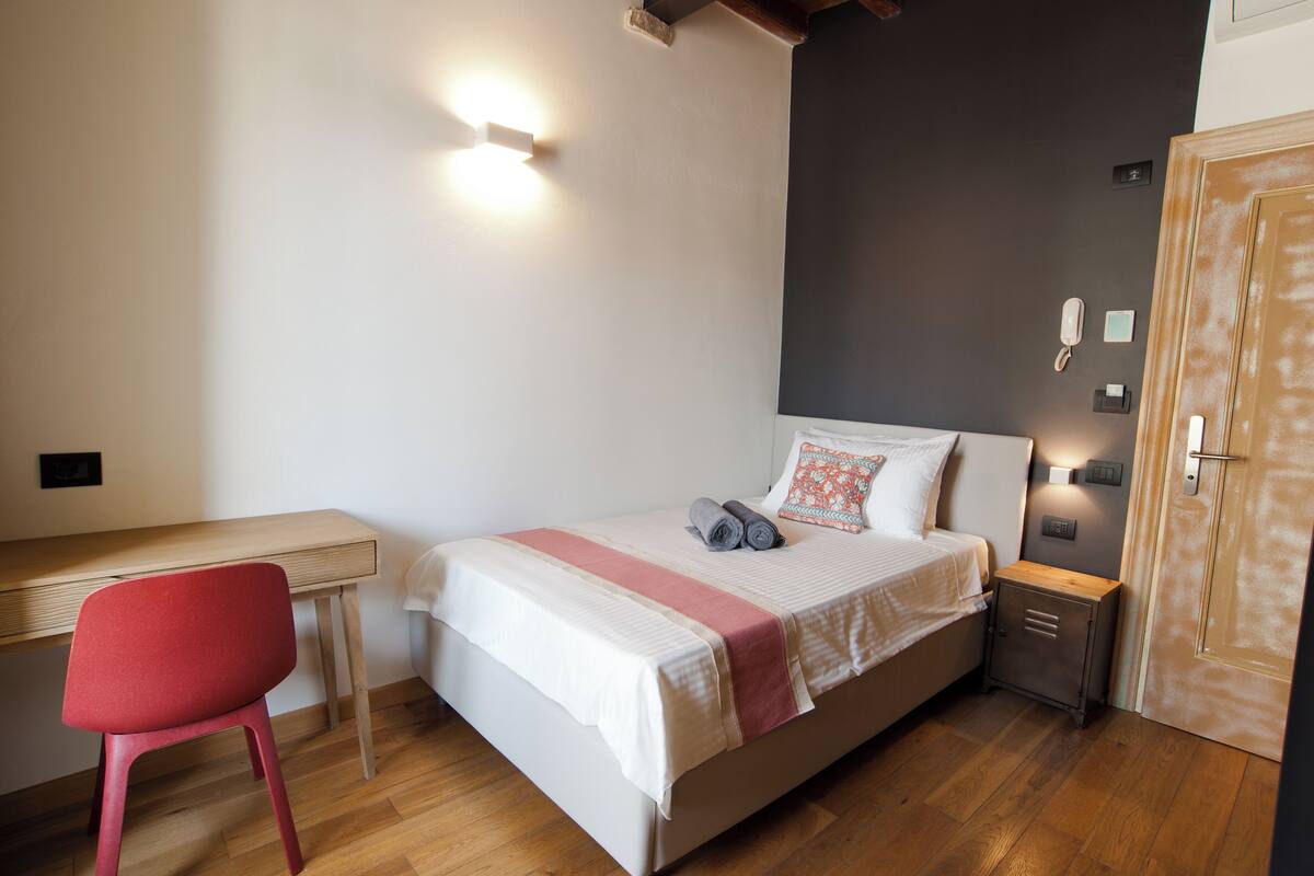 Quarto com uma cama, escrivaninha, cadeira, mesa de cabeceira e porta.