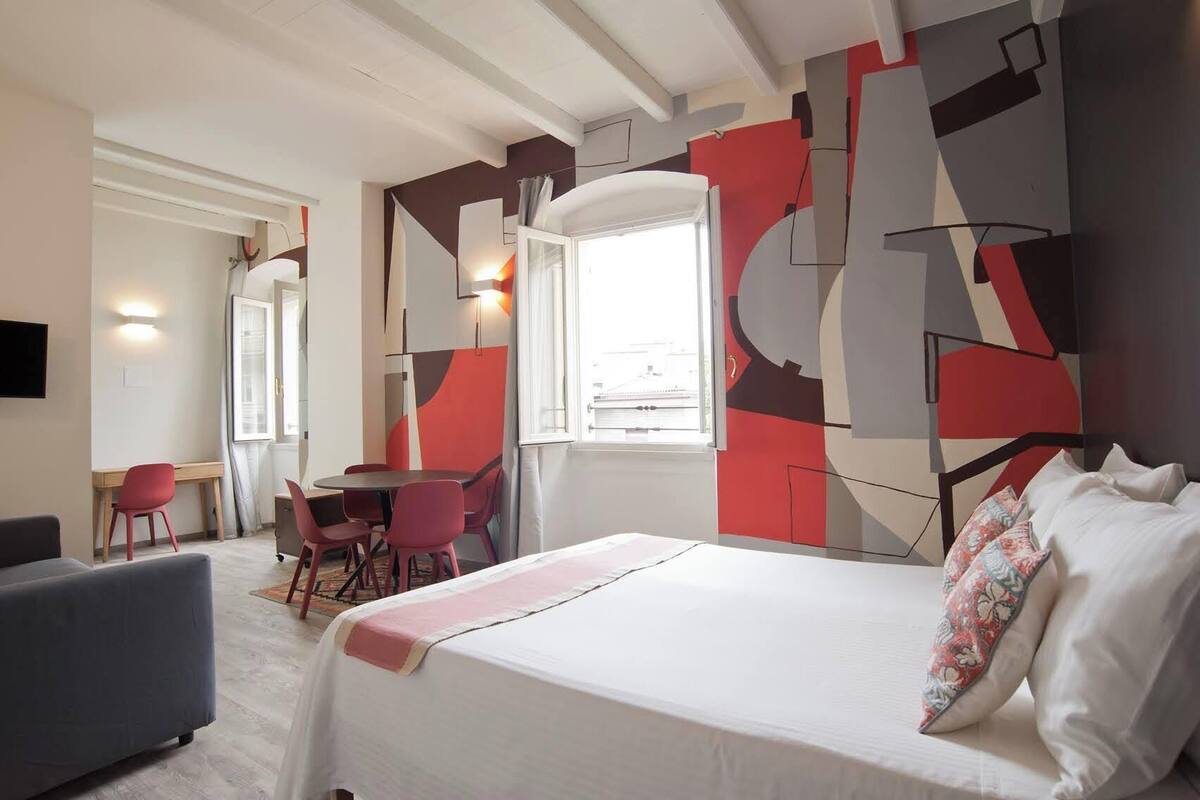 Quarto de hotel moderno com cama, secretária e cadeiras, além de um grande mural abstrato na parede.